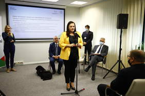 Pokaż zdjęcie: Wygłaszająca wystąpienie w sali konferencyjnej kobieta. Za nią tłumaczka języka migowego i mężczyzna z psem przewodnikiem. Wokół zgromadzeni słuchacze.