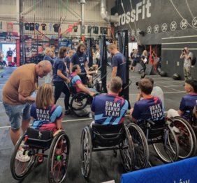 Pokaż zdjęcie: Sportowcy z niepełnosprawnościami na wózkach inwalidzkich, biorący udział w zawodach w przestrzeni sportowej. Po lewej stronie nachyla się do nich stojący mężczyzna w jasnobrązowej koszulce. Na drugim planie sportowiec na wózku inwalidzkim w otoczeniu trenerów. Otoczenie to duża hala sportowa przeznaczona do zawodów typu cross fit.