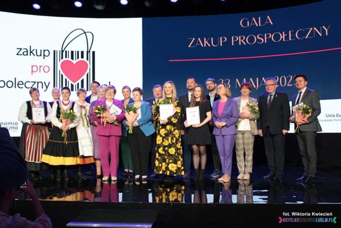 Pokaż zdjęcie: Na scenie stoją laureaci konkursu Zakup Prospołeczny, trzymają w rękach przyznane certyfikaty. Na scenie od lewej stoją również artyści w strojach ludowych.