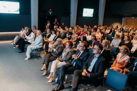 Widok na scenę podczas konferencji, sfotografowany z perspektywy publiczności siedzącej w pierwszych rzędach. Po lewej stronie sceny, przy mównicy z herbem Szczecina (Gryfem), stoi i przemawia kobieta w jasnej marynarce. Po prawej stronie ekranu stoi druga kobieta w niebieskiej sukience, trzymająca w dłoniach dokumenty. W tle widoczny jest duży ekran wyświetlający grafikę z napisem „Ogólnopolska Konferencja »Codzienność w spektrum relacji. Niewidoczni bohaterowie«”. Za prelegentkami wyeksponowano czarno-białe fotografie na sztalugach.