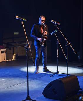 Pokaż zdjęcie: Mężczyzna w ciemnym garniturze, różowym krawacie i okularach przeciwsłonecznych stoi na środku sceny i gra na saksofonie. Artysta jest oświetlony jasnym punktowym reflektorem, który tworzy biały krąg na podłodze. Przed nim ustawione są trzy statywy z mikrofonami. Tło jest ciemne, minimalistyczne, z widocznym w głębi znakiem wyjścia ewakuacyjnego.