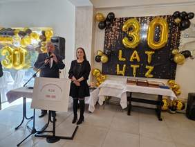 Pokaż zdjęcie: Kobieta i mężczyzna przemawiają do mikrofonu. W tle na ścianie widoczny napis z balonów 30 lat WTZ.