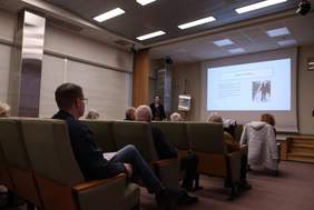 Pokaż zdjęcie: Na zdjęciu widać salę konferencyjną. Na fotelach siedzą ludzie słuchający wykładu prelegentki. Pośrodku pomieszczenia znajduje się duży ekran, na którym wyświetlana jest prezentacja.