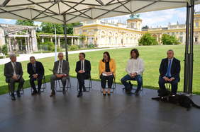 Pokaż zdjęcie: Grupa osób siedząca na krzesłach, w tle trawnik i zabudowania pałacowe