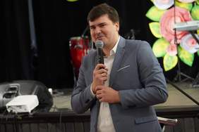 Pokaż zdjęcie: Mężczyzna w szarej marynarce i białej koszuli trzyma mikrofon, za nim scena z instrumentami