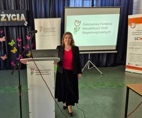 Pokaż zdjęcie: Jedna z prelegentek, uśmiechnięta kobieta z długimi włosami i czerwonej bluzce, pozuje do zdjęcia.