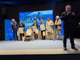 Pokaż zdjęcie: Grupa siedmiu osób na scenie podczas oficjalnej uroczystości. Pięć osób trzyma duże, oprawione w ramy dyplomy oraz białe torby upominkowe. Na dyplomach widoczne są kwoty (np. 8 000 PLN, 9 000 PLN). W centrum znajduje się kobieta na wózku inwalidzkim. Po prawej stronie, na pierwszym planie, stoi mężczyzna w okularach wykonujący gesty rękami (prawdopodobnie tłumacz języka migowego). W tle ekran z grafiką przedstawiającą pływaka oraz napis „SPORTOWE POMORZE ZACHODNIE”.