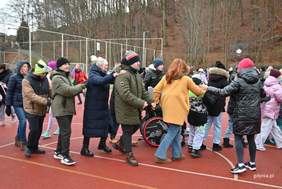 Pokaż zdjęcie: Grupa osób na zewnątrz, na bieżni sportowej, trzyma się za ramiona i tańczy w długim wężyku. Wśród uczestników są osoby w zimowych kurtkach oraz osoba na wózku. W tle las bez liści i ogrodzenie boiska. Atmosfera radosna, integracyjna.
