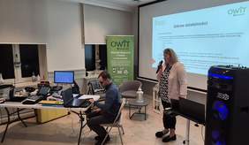 Pokaż zdjęcie: Na zdjęciu widzimy prezentację organizowaną przez OWiT Szczecin – Ośrodek Wsparcia i Testów. Wydarzenie odbywa się w eleganckim, jasnym pomieszczeniu z zasłoniętymi oknami. Na pierwszym planie po prawej stronie kobieta w jasnej marynarce mówi do mikrofonu, stojąc przy dużym ekranie, na którym wyświetlona jest prezentacja zatytułowana „Zakres działalności”. Obok niej stoi duży głośnik z niebieskim podświetleniem.  Po lewej stronie mężczyzna siedzi przy stole, pracując na laptopie. Na stole znajduje się wiele urządzeń elektronicznych, prawdopodobnie technologii wspomagających dla osób z niepełnosprawnościami (np. terminale, urządzenia brajlowskie, monitory z powiększonym tekstem). Za nim stoi baner informacyjny OWiT Szczecin, zawierający opis usług i dane kontaktowe.