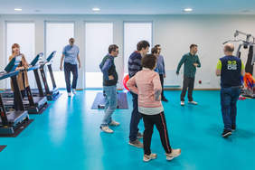 Pokaż zdjęcie: Grupa osób w sali treningowej z bieżniami, słucha instruktora. Wszyscy ubrani swobodnie, część stoi, inni się poruszają. W tle jasne okna, turkusowa podłoga i sprzęt do ćwiczeń.