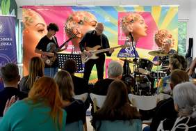 Muzycy występują przed publicznością podczas wydarzenia festiwalowego. Widać gitarzystę, basistę i perkusistę grających na scenie, za którą znajduje się kolorowa scenografia 23. Europejskiego Festiwalu Filmowego Integracja „Ja i Ty”.