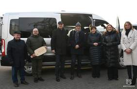 Pokaż zdjęcie: Grupa siedmiu osób stojących przed jasnoszarym busem. Wszyscy są ubrani w zimowe ubrania: płaszcze i kurtki, większość w ciemnych kolorach, jedna osoba po prawej w jasnym, beżowym płaszczu. Od lewej stoi mężczyzna w ciemnej kurtce i ciemnej czapce, obok niego osoba w pikowanej, oliwkowozielonej kurtce trzyma w dłoniach duży, jasny, drewniany „klucz” w kształcie deski. Bus w tle jest zaparkowany bokiem, ma duże, przyciemniane boczne okno i otwarte boczne drzwi. Na samym dole po prawej stronie fotografii znajduje się małe logo z napisem „KROTOSZYN”. 