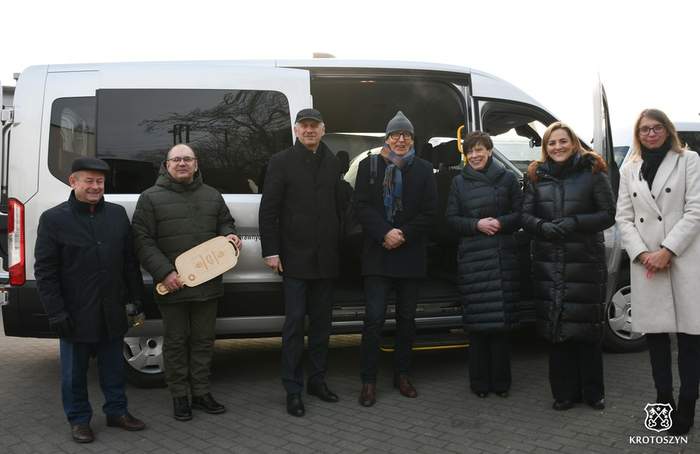 Pokaż zdjęcie: Grupa siedmiu osób stojących przed jasnoszarym busem. Wszyscy są ubrani w zimowe ubrania: płaszcze i kurtki, większość w ciemnych kolorach, jedna osoba po prawej w jasnym, beżowym płaszczu. Od lewej stoi mężczyzna w ciemnej kurtce i ciemnej czapce, obok niego osoba w pikowanej, oliwkowozielonej kurtce trzyma w dłoniach duży, jasny, drewniany „klucz” w kształcie deski. Bus w tle jest zaparkowany bokiem, ma duże, przyciemniane boczne okno i otwarte boczne drzwi. Na samym dole po prawej stronie fotografii znajduje się małe logo z napisem „KROTOSZYN”. 