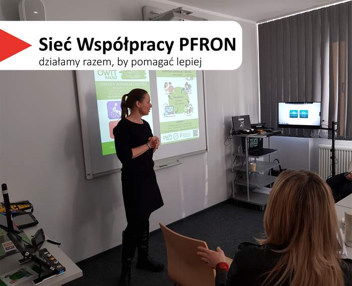 Pokaż zdjęcie: W centralnej części slajdu widać kobietę w czarnym stroju, stojącą przy tablicy projektowej i mówiącą do grupy. W tle jest wyświetlana prezentacja na tablicy. Po prawej stronie sali stoi wózek ze sprzętem i monitor, a przy oknie są zasłony. Na pierwszym planie widać siedzącą osobę z jasnymi włosami, od tyłu, zwróconą w stronę prowadzącej. Na slajdzie w lewym górnym rogu jest biały pasek z czerwonym trójkątem i dużym napisem: „Sieć Współpracy PFRON”, a pod nim mniejszy: „działamy razem, by pomagać lepiej”.