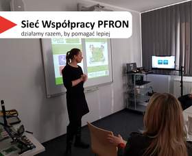 W centralnej części slajdu widać kobietę w czarnym stroju, stojącą przy tablicy projektowej i mówiącą do grupy. W tle jest wyświetlana prezentacja na tablicy. Po prawej stronie sali stoi wózek ze sprzętem i monitor, a przy oknie są zasłony. Na pierwszym planie widać siedzącą osobę z jasnymi włosami, od tyłu, zwróconą w stronę prowadzącej. Na slajdzie w lewym górnym rogu jest biały pasek z czerwonym trójkątem i dużym napisem: „Sieć Współpracy PFRON”, a pod nim mniejszy: „działamy razem, by pomagać lepiej”.