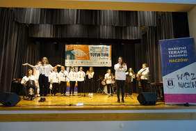 Występ sceniczny, grupa 12 osób w białych bluzach i ciemnych spodniach