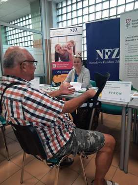 Pokaż zdjęcie: Starszy pan z kulą siedzi przy stole naprzeciwko pani. Kobieta uśmiecha się do niego. Na stole leżą różne poradniki, informatory, ulotki reklamowe. 