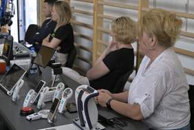 Pokaż zdjęcie: Na stole ustawiony jest asystujący sprzęt. Za stołem siedzą trzy kobiety i mężczyzna, zwróceni bokiem do widza, uważnie słuchają 