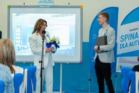 Prelegenci podczas konferencji stoją przy mikrofonie, w tle ekran z wyświetlonym slajdem