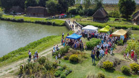 Pokaż zdjęcie: Zdjęcie z drona, pokazujące piknik w skansenie, jezioro, namioty, uczestników.