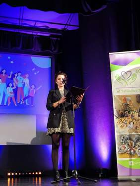 Pokaż zdjęcie: Na scenie kobieta ubrana w sukienkę czyta list gratulacyjny od pani dyrektor.