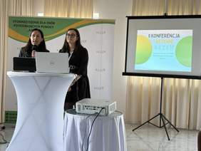 Pokaż zdjęcie: W sali stoją dwie prelegentki. Jedna z kobiet przemawia do mikrofonu. Przed paniami jest stolik, na którym leżą otwarte laptopy. 