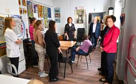 Pokaż zdjęcie: Grupa kilku osób stoi w sali udekorowanej pracami plastycznymi. Na jednej ze ścian wisi kalendarz.