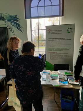 Pokaż zdjęcie: na zdjęciu dwie osoby stoją przy stoisku z materiałami reklamowymi PFRON w tle bilbord  PFRON 