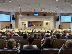 Pokaż zdjęcie: Na Sali konferencyjnej siedzi wiele osób , na dole sali mównica za którą stoi kobieta obok stół za który siedzą dwie osoby. Po obu stronach wielkie telebimy z prezentacjami. Na ścianie godło województwa pomorskiego.