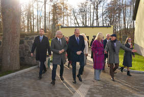 Ośmiu ludzi, głównie w płaszczach, idzie po chodniku z kostki brukowej w otoczeniu drzew bez liści i jasnego budynku. Słońce świeci zza drzew, rzucając jasne refleksy. Osoba idąca po prawej gestykuluje w przód.
