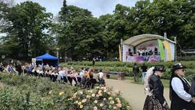 Pokaż zdjęcie: Widok na plenerową scenę w parku różanym, na której odbywa się występ artystyczny – grupa osób śpiewa lub przemawia. Przed sceną zebrana jest publiczność, siedząca na krzesłach, w tym osoby na wózkach inwalidzkich. W tle zielone drzewa, po lewej stronie niebieski namiot. Na pierwszym planie róże w pełnym rozkwicie. Po prawej stronie dwie kobiety w stylizacjach steampunkowych.