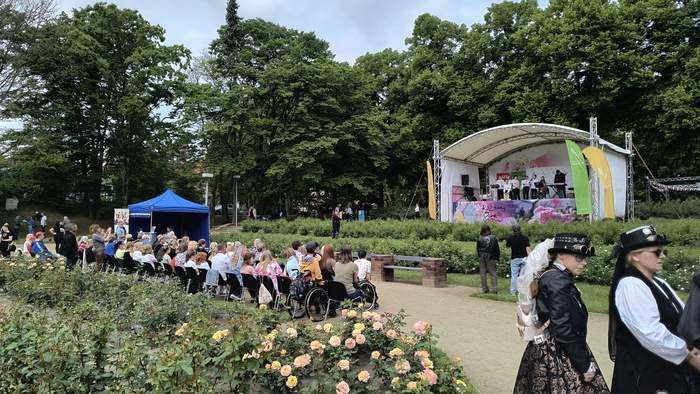 Pokaż zdjęcie: Widok na plenerową scenę w parku różanym, na której odbywa się występ artystyczny – grupa osób śpiewa lub przemawia. Przed sceną zebrana jest publiczność, siedząca na krzesłach, w tym osoby na wózkach inwalidzkich. W tle zielone drzewa, po lewej stronie niebieski namiot. Na pierwszym planie róże w pełnym rozkwicie. Po prawej stronie dwie kobiety w stylizacjach steampunkowych.