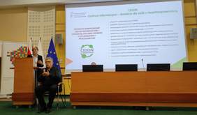 Pokaż zdjęcie: Na zdjęciu Edyta Kondzior stoi przy mównicy i wygłasza prelekcję podczas konferencji. Obok niej siedzi tłumacz języka migowego wykonujący przekład. W tle widoczny jest duży ekran z prezentacją dotyczącą usług informacyjno-doradczych CIDON oraz flagi Polski, Unii Europejskiej i województwa.