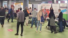 Tłum ludzi przechodzi obok stoisk informacyjnych. Na stoiskach są materiały informacyjne, za stoiskami widoczne logotypy wystawców.