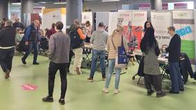 Pokaż zdjęcie: Tłum ludzi przechodzi obok stoisk informacyjnych. Na stoiskach są materiały informacyjne, za stoiskami widoczne logotypy wystawców.