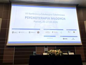 Ekran projekcyjny w sali konferencyjnej z wyświetloną planszą wydarzenia. Na środku slajdu jest duży tytuł, a pod nim data i miejsce. Na dole znajdują się logotypy partnerów. Pod ekranem stoi długi stół, a na nim w centrum kompozycja kwiatowa. Po bokach widać drobny sprzęt nagłośnieniowy. Tekst widoczny na slajdzie: VII Konferencja Edukacyjno-Szkoleniowa, a pod nim PSYCHOTERAPIA MŁODYCH oraz data i miejsce wydarzenia: Poznań, od 25 do 27 marca 2026 roku