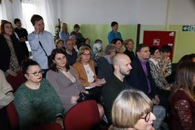 Pokaż zdjęcie: Zbliżenie na widownię, wśród gości siedzą elegancko ubrane kobiety i mężczyźni. Kilka osób stoi za osobami siedzącymi na krzesłach. Jedna z kobiet spogląda w stronę aparatu.