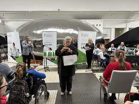 Pokaż zdjęcie: Starsza kobieta w siwych krótkich włosach i okularach ubrana w ciemny płaszcz trzyma w rękach statuetkę wykonaną w kształcie tulipana i torbę z logo PFRON.  Za nią mężczyzna trzymający mikrofon i banery z logo PFRON. Po bokach siedzą ludzie.