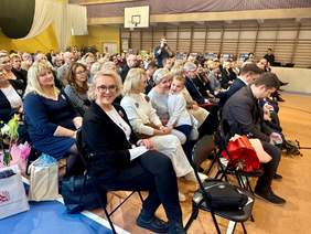 Pokaż zdjęcie: W szkolnej sali gimnastycznej siedzą na krzesłach w rzędach goście. Wszyscy są elegancko ubrani. Niektórzy trzymają w dłoniach kwiaty i upominki. 