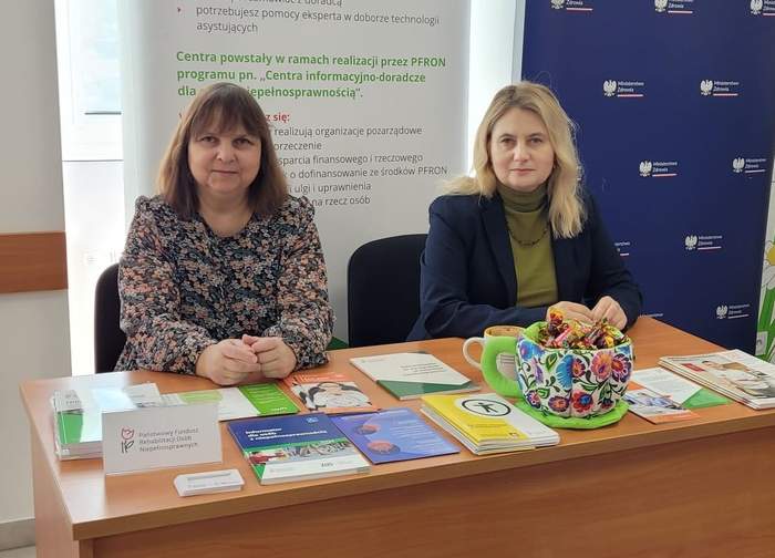Pokaż zdjęcie: Przy biuru siedzą dwie kobiety. Na stole leżą ulotki i informatory. W tle baner z logo PFRON.