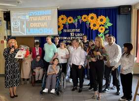 Pokaż zdjęcie: Grupa osób podczas jubileuszu 10-lecia Stowarzyszenia Otwarte Drzwi w Lęborku; kobieta z mikrofonem i bukietem kwiatów przemawia, obok niej uczestnicy wydarzenia, w tym osoby z niepełnosprawnościami. W tle dekoracja z napisem i słonecznikami.