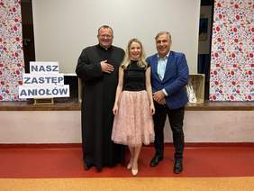 Pokaż zdjęcie: Na zdjęciu stoi Ksiądz w okularach, ubrany w czarną sutannę,  kobieta w blond włosach i rozłożystej kwiecistej sukience, mężczyzna w granatowej marynarce ciemnych spodniach i ciemnych butach. Obok drukowany napis NASZ ZASTĘP ANIOŁÓW. 