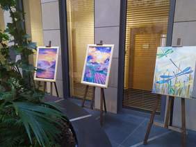 Pokaż zdjęcie: Na tle kamiennej ściany stoją sztalugi, a na nich są wyeksponowane trzy obrazy, po lewej stronie zdjęcia kompozycja z roślin zielonych