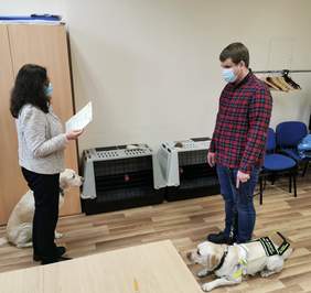 Pokaż zdjęcie: Stoją naprzeciwko siebie, w pewnej odległości, kobieta i mężczyzna. Kobieta trzyma kartkę papieru. Towarzyszą im psy.