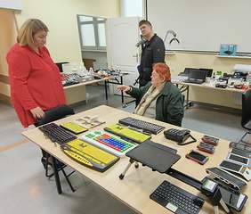 Pokaż zdjęcie: Kobieta w czerwonej bluzce rozmawia ze starszą kobietą siedzącą przy stole, na którym znajdują się klawiatury o powiększonych klawiszach i inne urządzenia technologiczne. W tle mężczyzna obserwuje rozmowę, a na stołach widoczne są różne sprzęty elektroniczne.