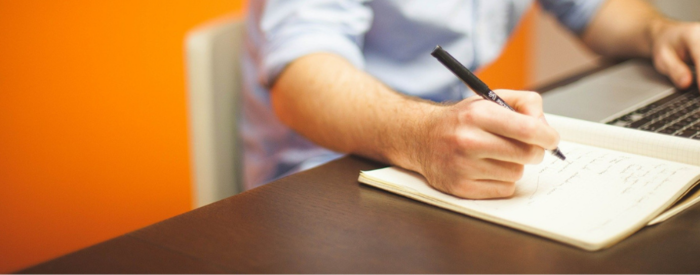 Mężczyzna siedzący przy biurku. W dłoni trzyma długopis, na biurku leży zeszyt z notatkami oraz klawiaturę komputera.