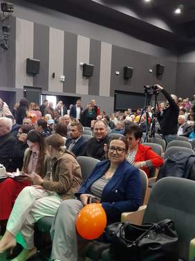 Pokaż zdjęcie: Sala kinowa, na fotelach siedzą zaproszeni goście. Kobieta na pierwszym planie trzyma pomarańczowy balon.