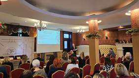 Pokaż zdjęcie: Na zdjęciu sala konferencyjna, w której odbywa się prezentacja dotycząca jubileuszu 10-lecia funkcjonowania Krajowego Funduszu Szkoleniowego. Widoczna jest grupa uczestników siedzących na czerwonych krzesłach, patrzących na mówcę stojącego przed ekranem projekcyjnym, na którym wyświetlana jest prezentacja. 