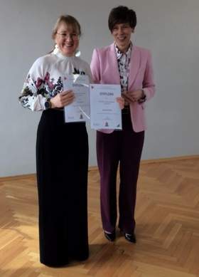 Dwie uśmiechnięte kobiety stoją przodem do aparatu w jasnym pomieszczeniu, trzymając w dłoniach dyplomy. Kobieta po lewej ma spięte jasne włosy z grzywką, okulary, białą bluzkę w kwiaty i długą czarną spódnicę. Kobieta po prawej ma krótkie ciemne włosy, różową marynarkę, wzorzystą koszulę i ciemne spodnie. Tło stanowi jasna ściana i drewniana podłoga.