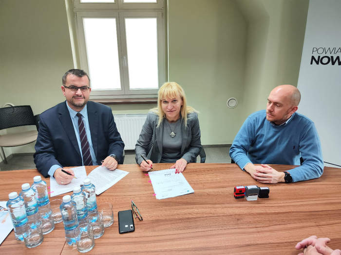 Pokaż zdjęcie: Przy stoliku w kolorze brązowym siedzą Dyrektor Andrzej Gonia, Starosta Iwona Brzozowska oraz przedstawiciel WTZ w Nowej Soli, wszyscy ubrani odświętnie, podpisują umowę